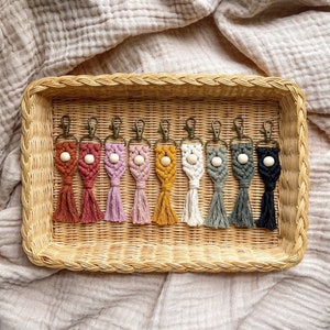 May include: A collection of handmade macrame keychains in various colors, including brown, pink, yellow, white, and black. Each keychain features a wooden bead and a tassel, displayed in a woven basket.
