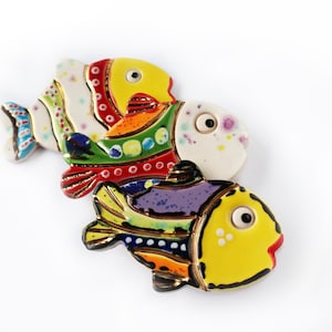 Puede incluir: Tres adornos de peces de cerámica con diseños abstractos coloridos. Los peces están apilados uno encima del otro, con el pez más grande en la parte inferior y el más pequeño en la parte superior. Los peces están pintados en una variedad de colores, incluyendo amarillo, rojo, verde, azul y morado.