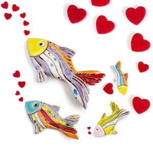 Puede incluir: Tres peces de cerámica coloridos con rayas rojas, amarillas, azules y moradas y detalles blancos. Los peces nadan hacia un rastro de corazones rojos.
