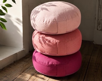 May include: A stack of three round, flat floor cushions in varying shades of pink. The top cushion is light pink, the middle is a medium rose pink, and the bottom cushion is a deep magenta. Each cushion features a side handle and zipper.