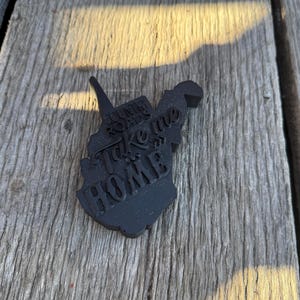 May include: Black West Virginia state outline with the text "Country Roads Take Me Home" in white lettering.