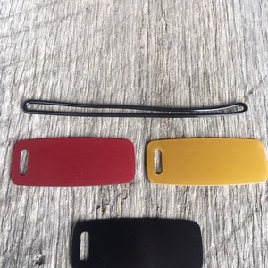 May include: A collection of small, rectangular tags in red, yellow, and black, each with a rounded end and a slot for attachment. A black, oval-shaped rubber band is above the tags, all resting on a weathered wooden surface.