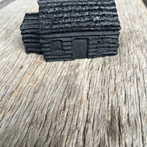 May include: A miniature black log cabin with a textured roof and visible door and window details. The cabin is made of stacked, dark-colored logs, and sits on a weathered wooden surface.
