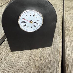 May include: A black wooden clock with a white face and silver hands. The clock is shaped like a small arch and is sitting on a wooden surface.