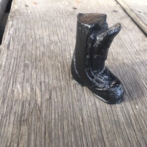 May include: A small black boot sculpture made of wood. The boot is detailed with a textured surface and a pointed toe.