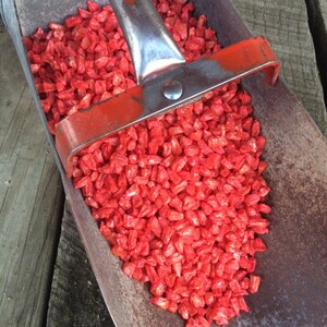 May include: A red metal scoop filled with bright red corn kernels. The scoop is made of metal and has a wooden handle.