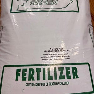 May include: A white bag of Kentucky Green fertilizer. The bag has green text that reads "FERTILIZER" and "CAUTION: KEEP OUT OF REACH OF CHILDREN." The bag also has the numbers "10-20-10" printed on it.