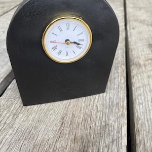 May include: A black clock with a gold rim and white face. The clock is sitting on a wooden surface.