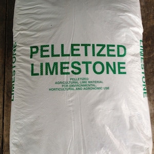 May include: A white plastic bag filled with white pelletized limestone. The bag has green text that reads "PELLETIZED LIMESTONE AGRICULTURAL LIME MATERIAL FOR ENVIRONMENTAL, HORTICULTURAL AND AGRONOMIC USE".