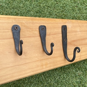 May include: Three black metal hooks with a decorative swirl at the end are mounted on a wooden board. The hooks are screwed into the board.