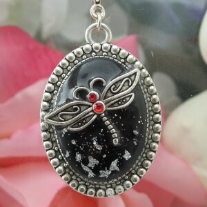 Puede incluir: Un colgante de plata con un diseño de libélula negro y plateado. La libélula tiene piedras de imitación rojas para los ojos y está colocada sobre un fondo negro con purpurina plateada.