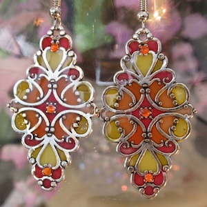 Puede incluir: Un par de pendientes de plata con un diseño de vitral. Los pendientes presentan una combinación de colores amarillo, naranja y rojo con pequeñas piedras preciosas rojas.