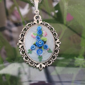 Puede incluir: Un collar con un colgante ovalado de plata con un diseño floral azul y blanco. El colgante presenta un grupo de flores azules con hojas verdes y acentos rosados.