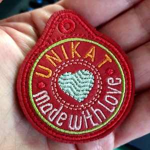 Puede incluir: Llavero de cuero rojo con un corazón verde y el texto "Unikat Made with love" en bordado amarillo y rosa.