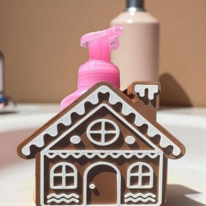 Könnte beinhalten: Ein braun-weißes Seifenspenderhalter in Lebkuchenhausform mit einem rosa Seifenspender. Der Halter hat ein detailliertes Design mit Fenstern, einer Tür und einem Schornstein. Der Spender ist über dem Dach sichtbar.