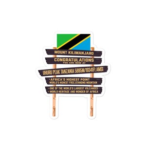 Op de afbeelding: Een houten bord met de tekst "Mount Kilimanjaro Congratulations You Are Now At Uhuru Peak Tanzania 5895m/19341ft AMSI" en een Tanzaniaanse vlag. Het bord bevat ook de tekst "Africa's Highest Point World's Highest Free-Standing Mountain One of the World's Largest Volcanoes World Heritage and Wonder of Africa."