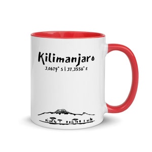May include: White ceramic mug with a red rim and handle. The mug features a black illustration of Mount Kilimanjaro and the text "Kilimanjaro 3.0674° S 37.3556° E".