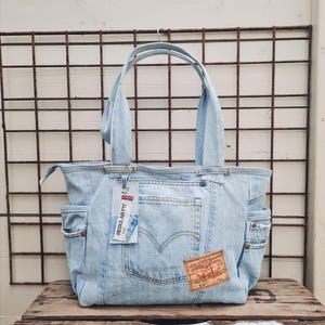 Könnte beinhalten: Hellblaue Jeans-Tragetasche mit zwei Außentaschen und einem Reißverschluss. Mit einem Levi Strauss & Co. Patch und einem "Regular Fit" Etikett. Die Tasche hat zwei Griffe und besteht aus recyceltem Denim.