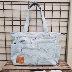 Op de afbeelding: Lichtblauwe denim tote bag met meerdere zakken en handvatten. De tas is gemaakt van hergebruikte denim jeans, met de originele zakken en een bruin leren label. De tas staat op een houten oppervlak.