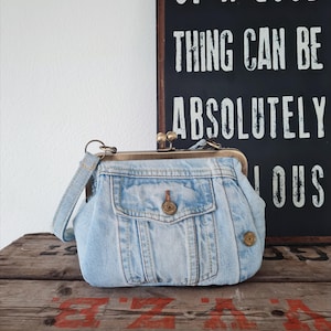 Peut inclure: Un sac à main en jean bleu clair avec une fermeture à fermoir en métal et une petite poche. Le sac a une sangle et une fermeture à bouton sur la poche. Une enseigne en arrière-plan indique "OF A GOOD THING CAN BE ABSOLUTELY LOUS".