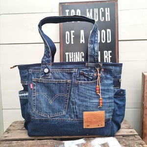 May include: A denim tote bag made from recycled jeans. The bag features multiple pockets, a leather patch, and a beaded charm. The text "TOO MUCH OF A GOOD THING" is visible in the background. The bag has a zipper closure and sturdy handles.