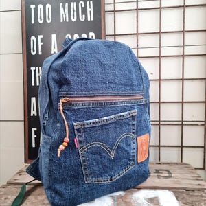 May include: A blue denim backpack with a front pocket, zipper, and a brown leather patch. The backpack has a brown zipper and a small leather tag with a string of beads. The text on the sign says "TOO MUCH OF A GOOD THING".