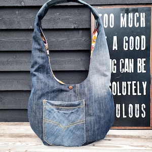 Op de afbeelding: Een hobo-stijl schoudertas gemaakt van blauwe denimstof, voorzien van een enkele brede schouderband, een voorzak met gele contraststiksels, een koperkleurige drukknoop in het midden en een zichtbare binnenvoering met een bloemenpatroon.