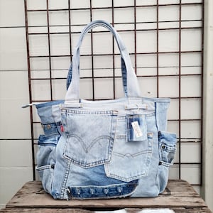 Op de afbeelding: Een lichtblauwe denim tote bag met meerdere zakken en een ritssluiting. De tas is gemaakt van hergebruikte denim en heeft het Levi's merklogo op een zak. De tas heeft twee handvatten en een label.
