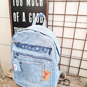May include: Light blue denim backpack with a front pocket, zipper, and a small tag. The backpack has a top handle and adjustable straps. The text "TOO MUCH OF A GOOD THING" is visible in the background.