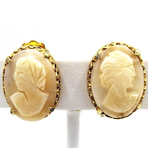 Può includere: Un paio di orecchini vintage a clip con cammeo. Ogni orecchino presenta un profilo color crema incastonato in una cornice ovale dorata con un bordo strutturato. Gli orecchini sono esposti su un supporto bianco su sfondo bianco.