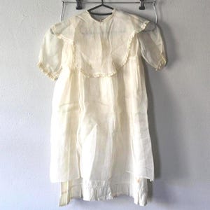May include: A vintage, cream-colored dress with short sleeves and a ruffled collar. The dress hangs on a metal hanger against a plain wall. The fabric appears lightweight and slightly sheer, with a layered skirt and delicate trim.