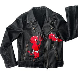 Puede incluir: Chaqueta de motorista negra de cuero sintético con cremallera asimétrica. Presenta gráficos de dibujos animados de diablitos rojos en la parte delantera. Cuello con solapas, bolsillos con cremallera y herrajes plateados.