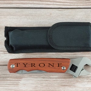 Puede incluir: Una multiherramienta con mango de madera grabado con "TYRONE" y cabeza de llave inglesa. Acompañada de una funda de transporte negra. La herramienta está diseñada para diversas tareas y es adecuada para proyectos de bricolaje.