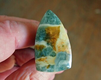 May include: A green, white, and brown cabochon gemstone with a pointed shape. The gemstone has a swirling pattern of colors.