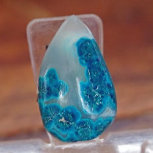 May include: A teardrop-shaped gemstone with a translucent, pale blue base and vibrant, deep turquoise patterns. The stone is set in a clear plastic holder, showcasing its natural beauty. The gemstone's unique color and design make it a striking piece.