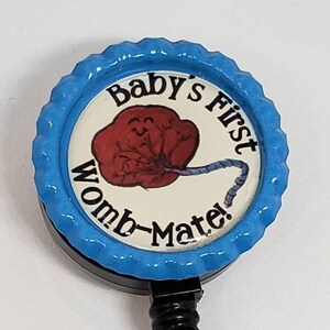 May include: Blue and white baby's first womb-mate badge reel with a cartoon image of a fetus and umbilical cord. The badge reel has a black retractable cord and a clear plastic badge holder.