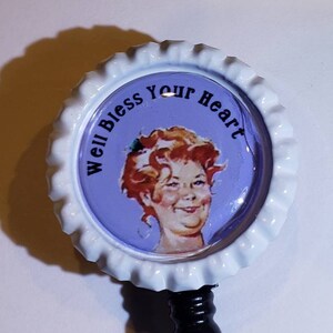 May include: A white plastic badge holder with a retractable cord and a round, white bottle cap with a purple background. The bottle cap has a picture of a woman with red hair and the text "Well Bless Your Heart".