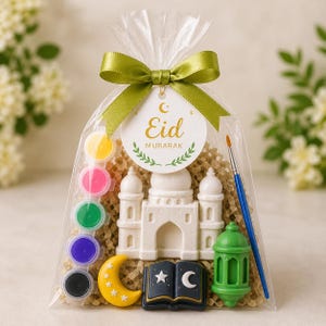 May include: A clear cellophane bag tied with a green ribbon, containing a white mosque-shaped item, paint pots, a paintbrush, a green lantern, a crescent moon, and a book with a star and crescent. The tag reads "Eid Mubarak."