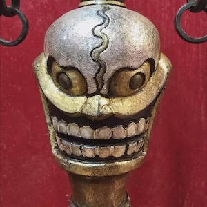 Puede incluir: Escultura ornamentada de una cara sonriente, predominantemente dorada y plateada, sobre un fondo rojo. La cara presenta ojos y dientes grandes. La escultura tiene una parte superior decorativa con anillos colgantes y una base detallada.