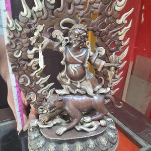 May include: A bronze and silver statue of a deity with a fierce expression, sitting on a tiger and surrounded by flames. The statue is intricately detailed and has a base with a decorative border.