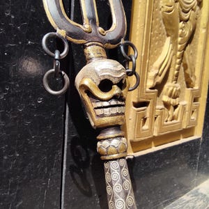 Puede incluir: Primer plano de un tridente decorativo con un mango en forma de calavera. Las púas del tridente son plateadas y doradas, con una cara de calavera y un mango estampado. Dos anillos metálicos cuelgan del tridente. El fondo es oscuro.