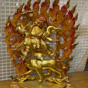 May include: A gold-colored statue of a deity with a fierce expression, riding a lion with flames behind them. The statue is on a circular base with intricate details.