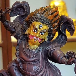 May include: A detailed sculpture of a deity with a fierce expression. The figure has a yellow face with three eyes, a red mouth, and orange and gold hair. It wears a dark robe and holds a weapon. The sculpture is likely of religious or cultural significance.