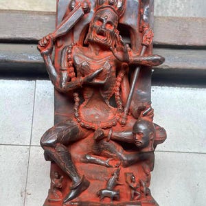 Può includere: Statua rossa e grigia raffigurante una figura seduta con più braccia, che tiene in mano armi e una testa mozzata. La statua presenta dettagli intricati ed è montata su una base rettangolare. La figura è adornata da una collana di perline.