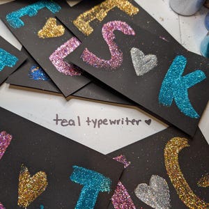 May include: Black rectangular cards with glitter letters and hearts in teal, gold, pink, and silver. The cards spell out the word "FSJK". The text "teal typewriter" is written in the center of the image.