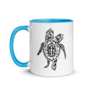 May include: A white ceramic mug with a light blue rim and handle. The mug features a black and white illustration of a two-headed sea turtle.
