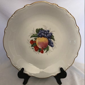 May include: White ceramic serving bowl with a scalloped edge and gold trim. The bowl features a central fruit design with an apple, grapes, and strawberries. The bowl is displayed on a black stand.