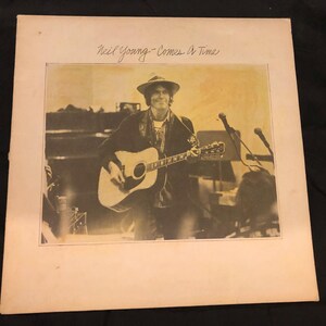 Puede incluir: Una portada de álbum de vinilo vintage con una foto en blanco y negro de Neil Young tocando la guitarra. El título del álbum "Comes A Time" está escrito en cursiva sobre la foto.