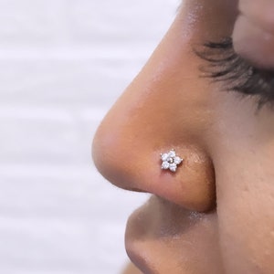 Puede incluir: Primer plano de la nariz de una persona con un pequeño anillo de nariz de plata en forma de flor con piedras transparentes.