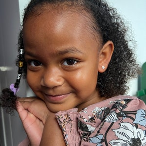 May include: A young girl with curly dark brown hair wearing a pink floral dress and small silver earrings. She is smiling and looking at the camera.